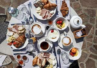 Vista dall'alto di un tavolo per la colazione sulla terrazza del Sunstar Hotel Piemonte: croissant, affettati, formaggi, frutta fresca, cappuccino e tè su un tavolo con tovaglia bianca